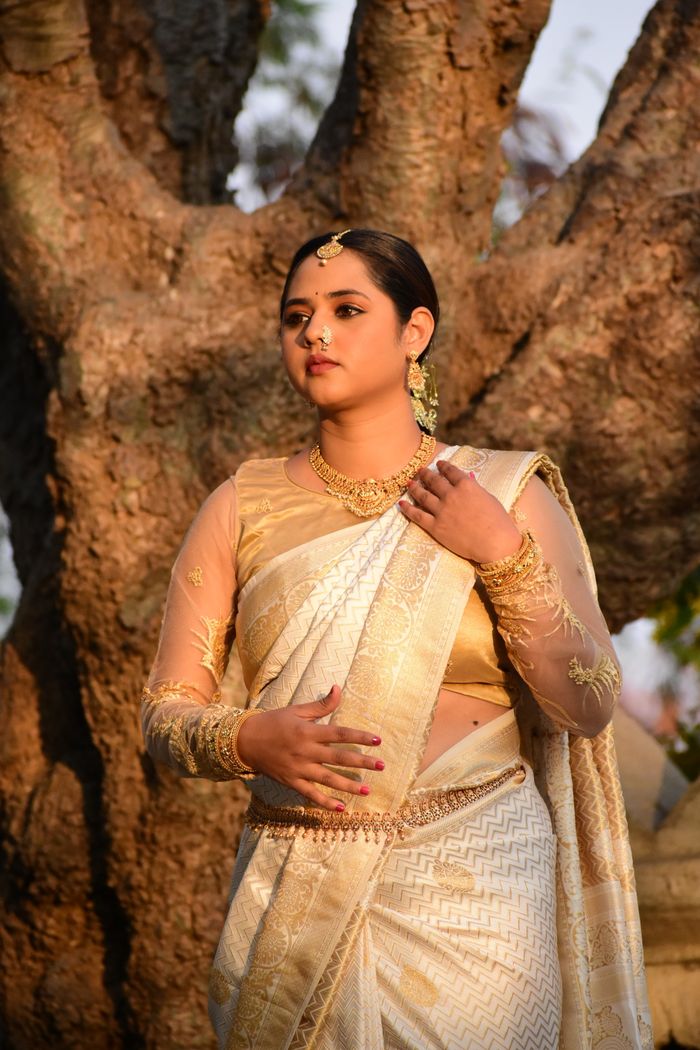 our-experience A woman in a golden saree stands gracefully by a tree, exuding cultural elegance.